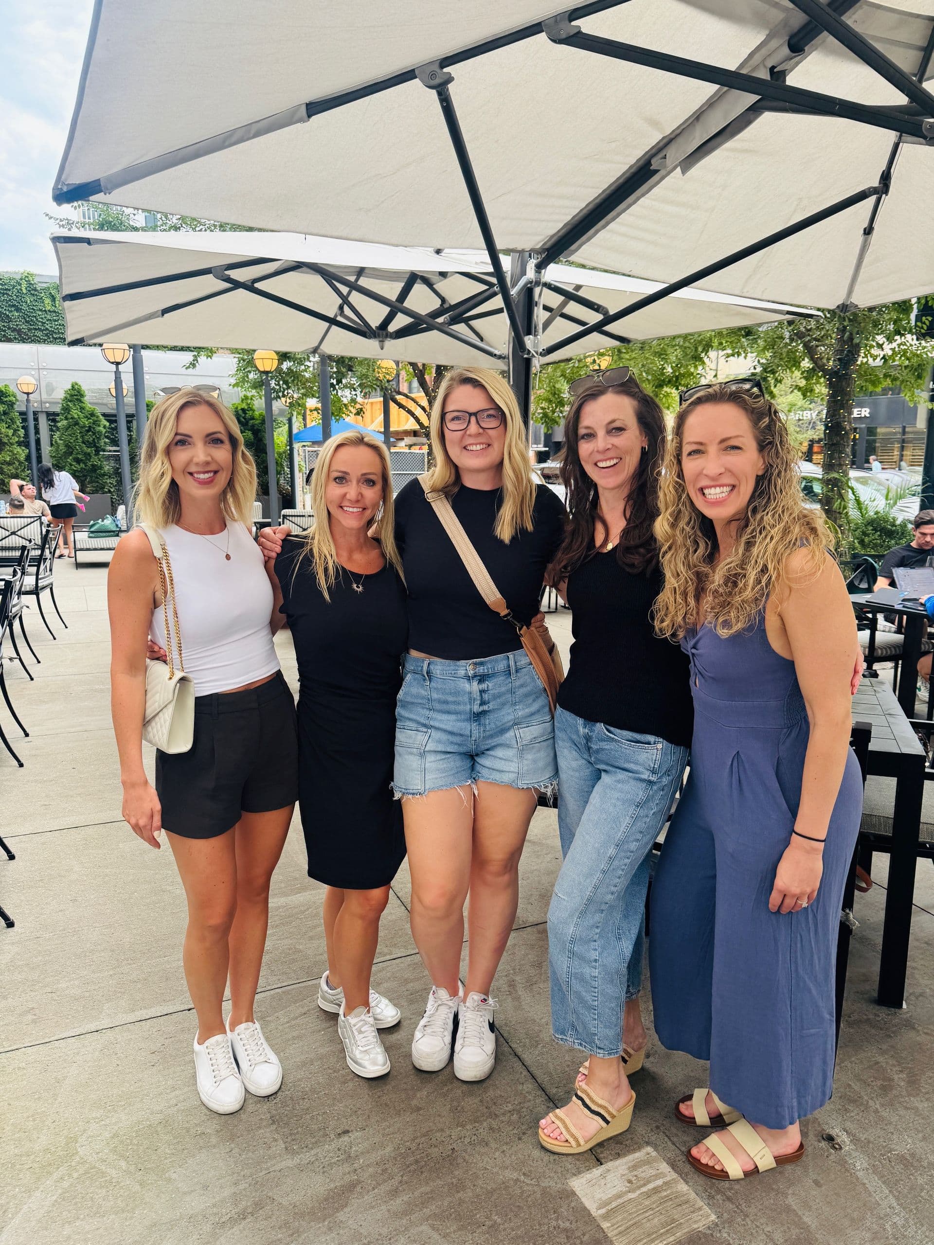 Five women smiling at an outdoor patio gathering under umbrellas, surrounded by trees and seating areas