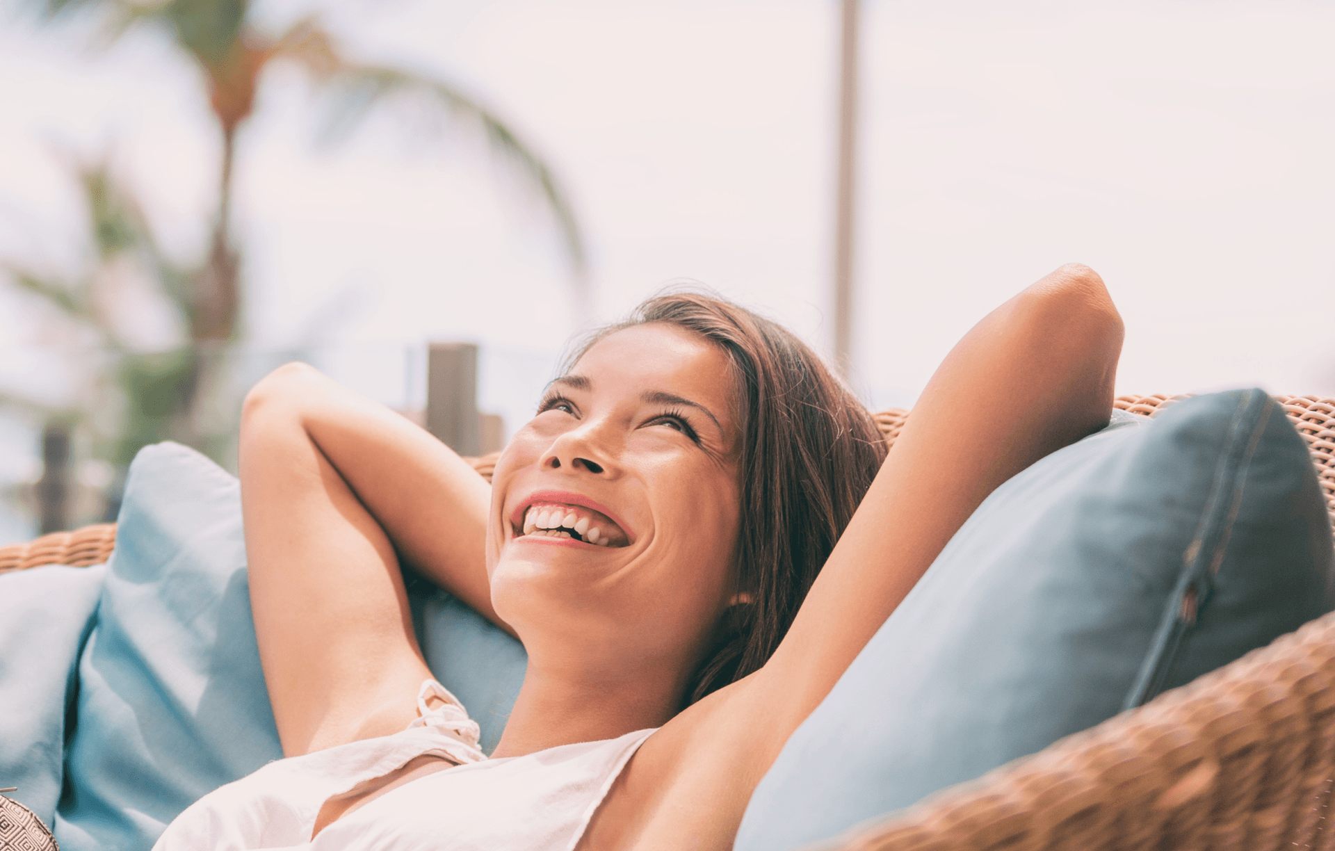 Woman relaxing on a wicker chair with arms behind head, smiling contentedly in a tropical setting with palm trees in the background