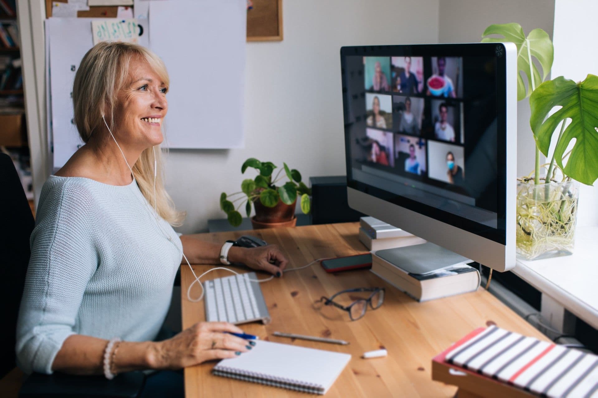 Woman in light blue sweater participating in video conference call with multiple participants displayed on monitor, workspace includes keyboard, plants, and notebooks