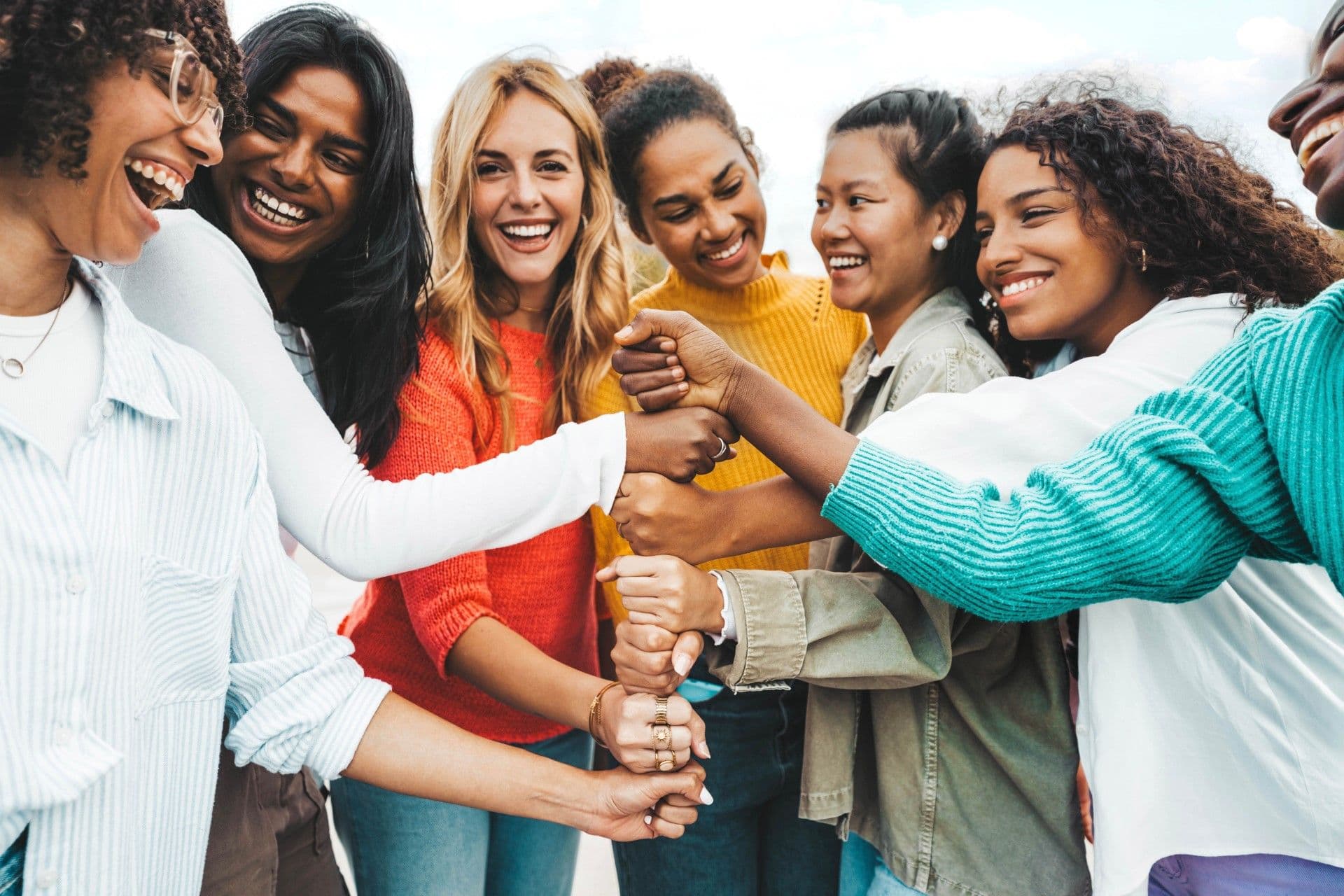 Group of diverse women smiling while stacking hands together in a circle, showing unity and teamwork outdoors