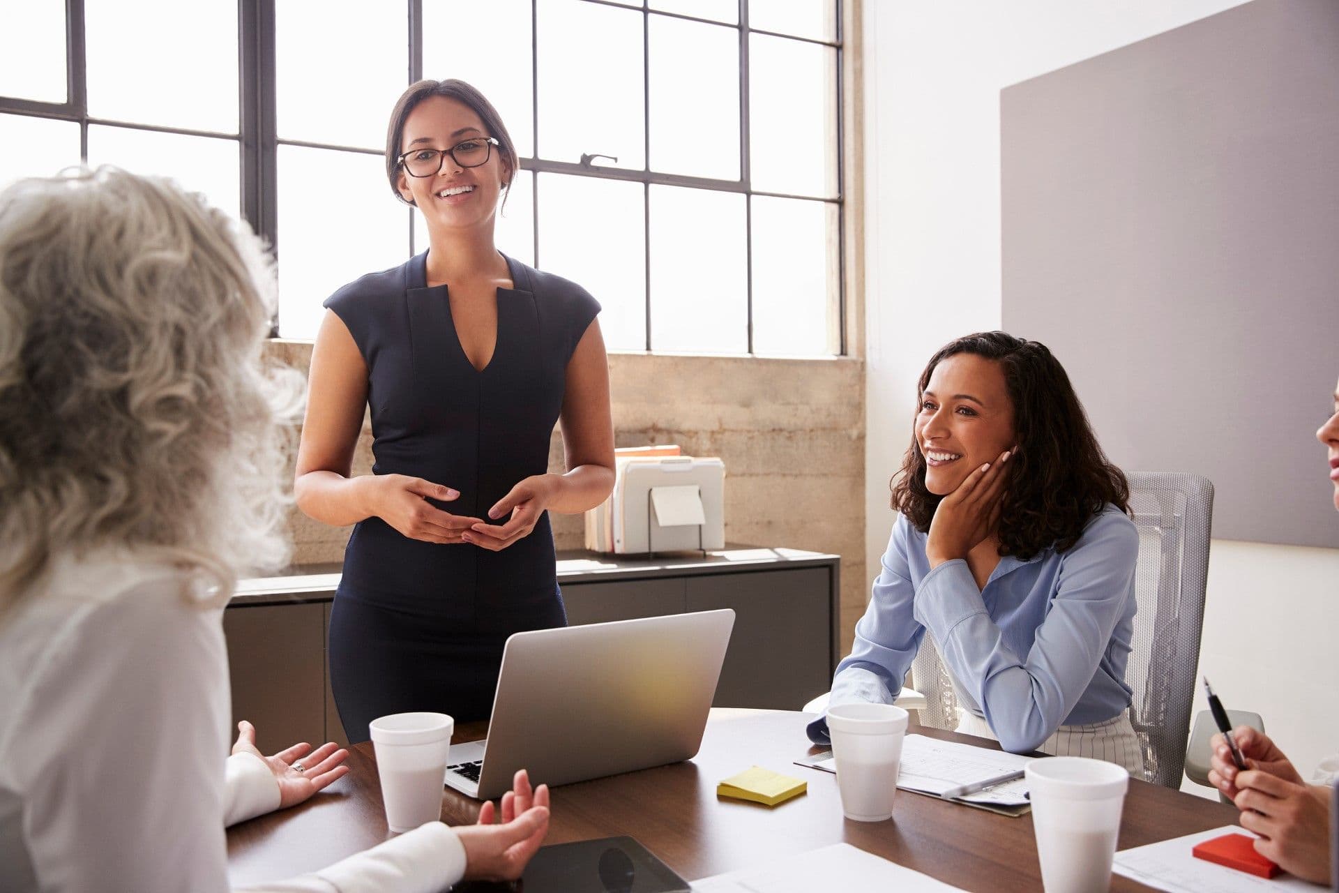Woman in black top and glasses leads a business meeting with colleagues seated at a table with laptop and coffee cups in a modern office