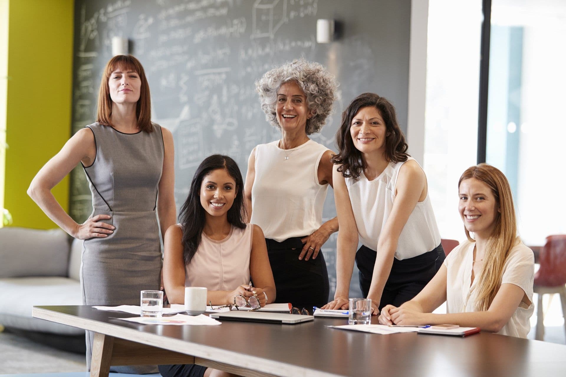 Diverse team of five professional women posing together at a conference table in a modern office with whiteboard in background