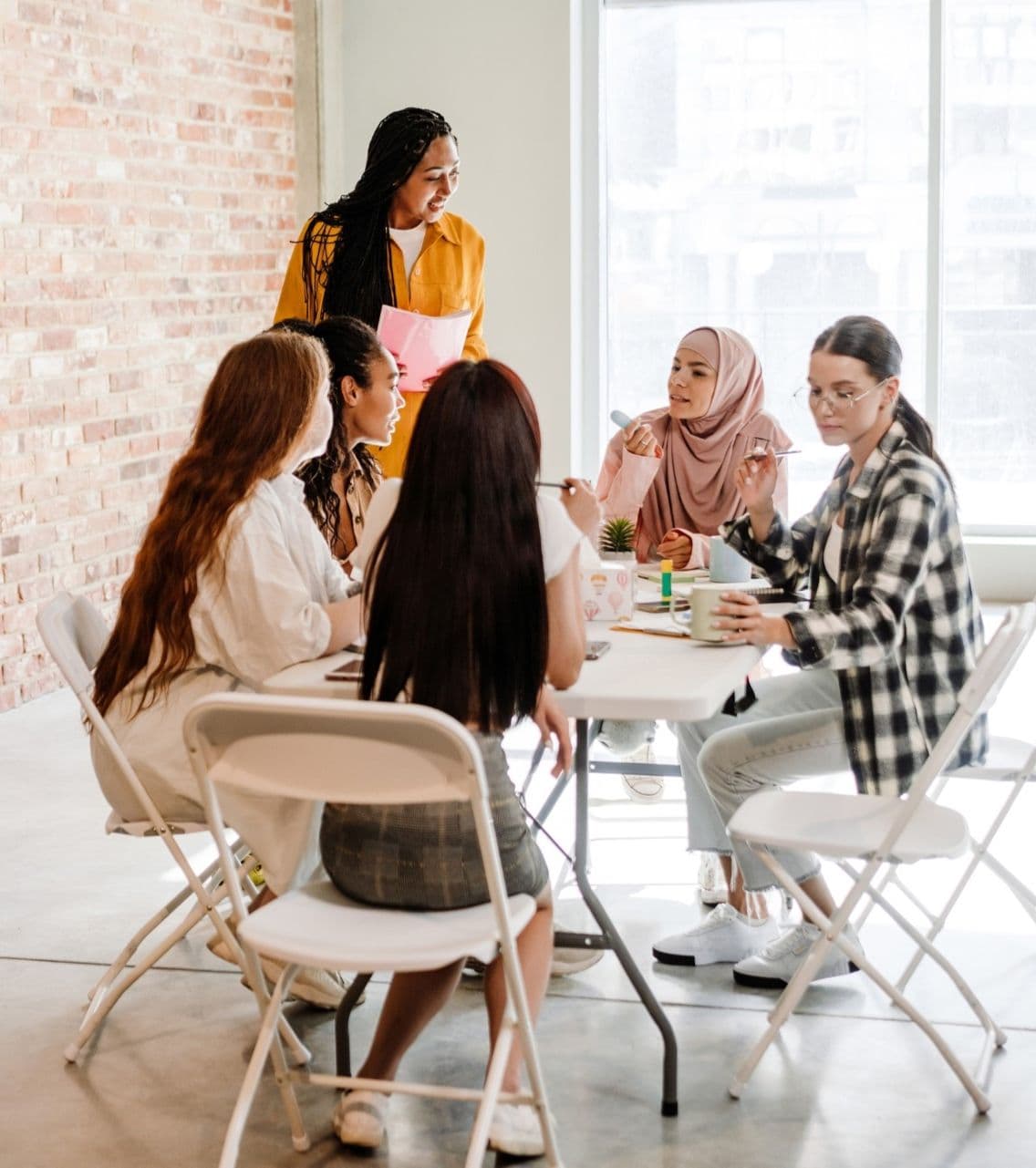 Group of five women in diverse attire meeting in a bright office space with exposed brick walls, sitting and standing around a table for a discussion or collaborative work session