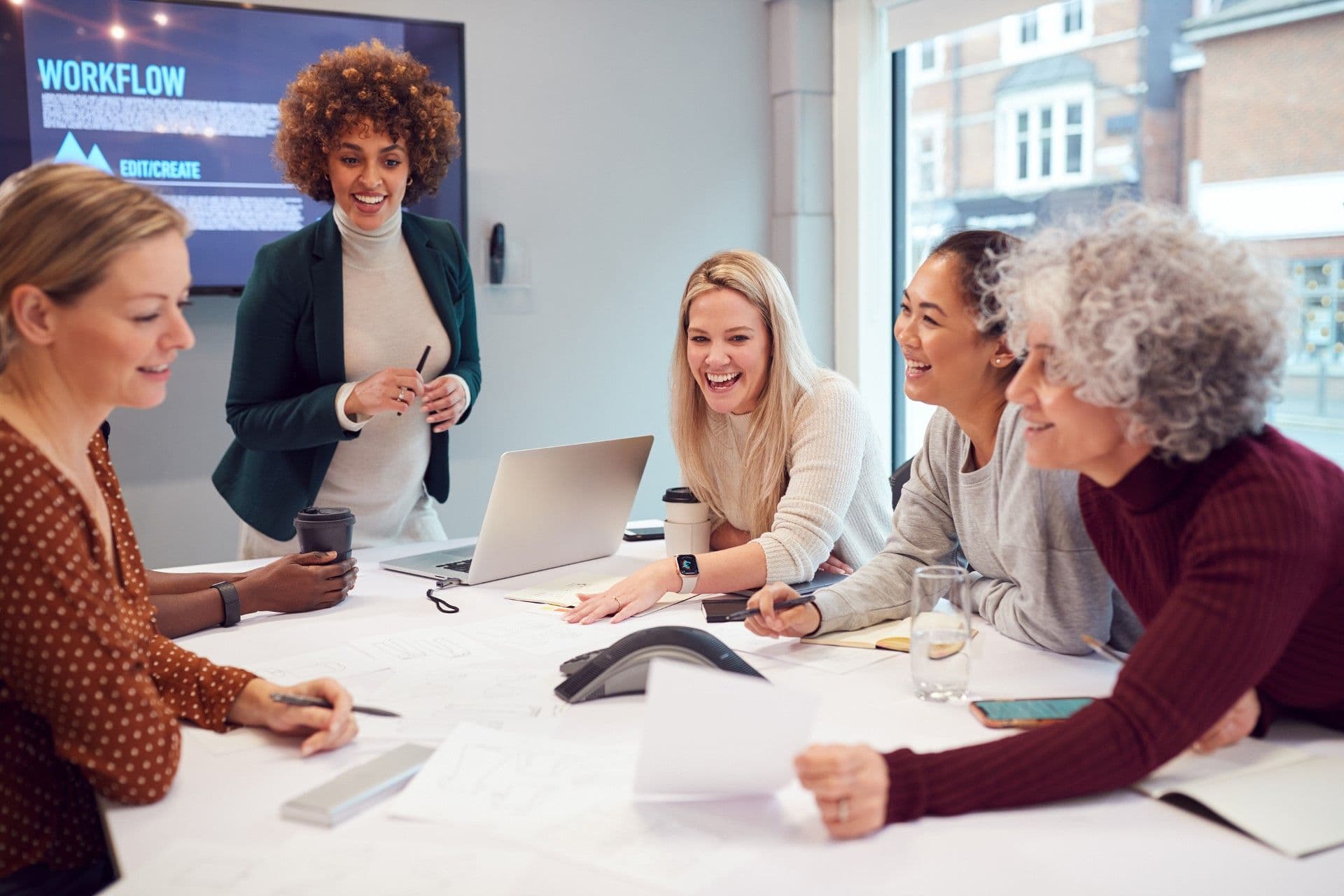 Diverse team of five women in a modern office meeting room gathered around a table with a laptop, smiling during a presentation with workflow diagram displayed on screen behind them