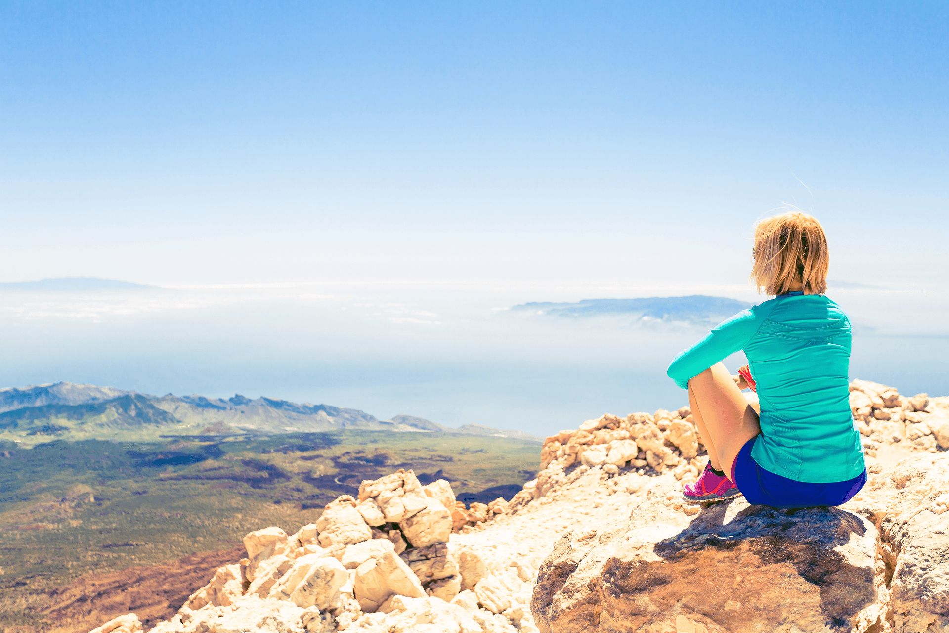 bigstock-Woman-Meditation-Beautiful-Ins-91037264 2.jpg Bright, wide-angle outdoor view of a female hiker wearing a vibrant teal long-sleeve athletic top and blue shorts, sitting on a rocky mountain summit and taking in panoramic views of distant islands surrounded by sea mist under a clear blue sky.