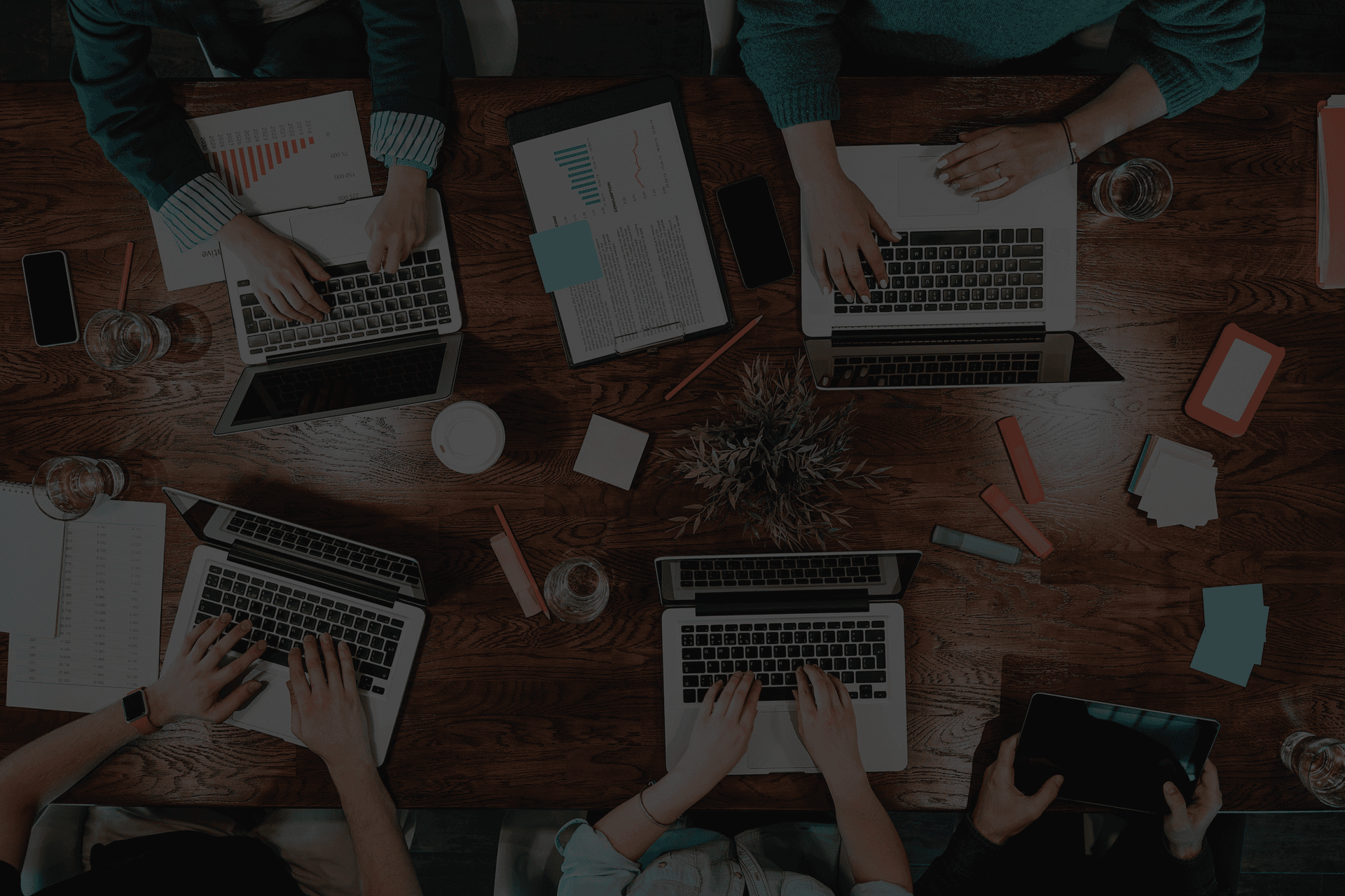 Dark overhead view of a collaborative group workspace where team members are actively typing on laptops, taking notes on clipboards and sticky notes, and using tablets around a rich dark wood table scattered with office supplies and highlighters.