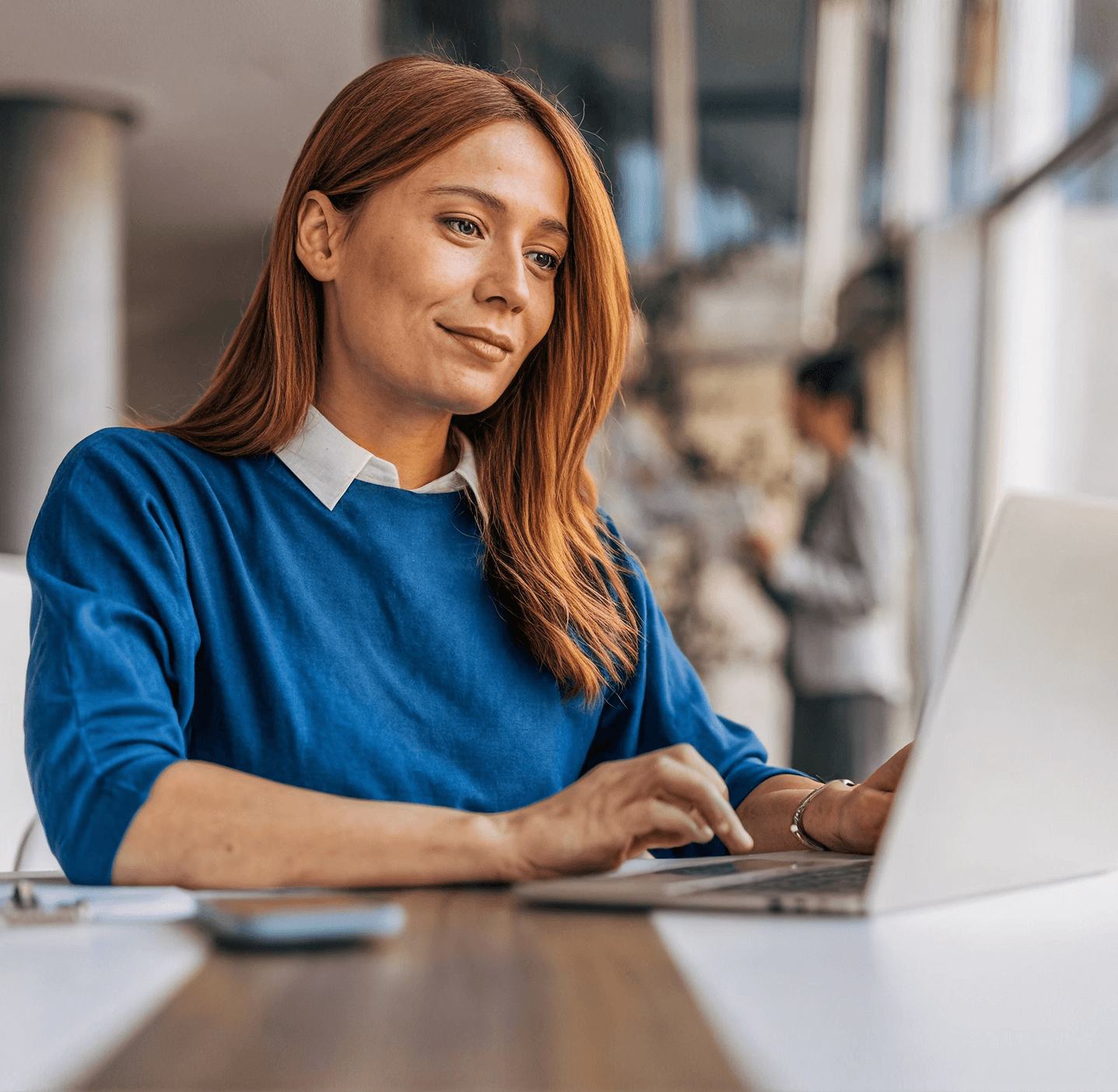 Woman with reddish-brown hair wearing blue sweater working on laptop at desk in office setting